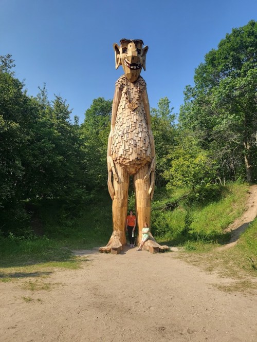 A large wooden troll sculpture towers over two people sitting on the ground in a lush green forest.