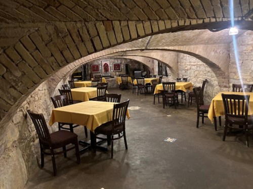 A cozy underground restaurant with arched brick ceilings, wooden chairs, and yellow tablecloths on round tables.