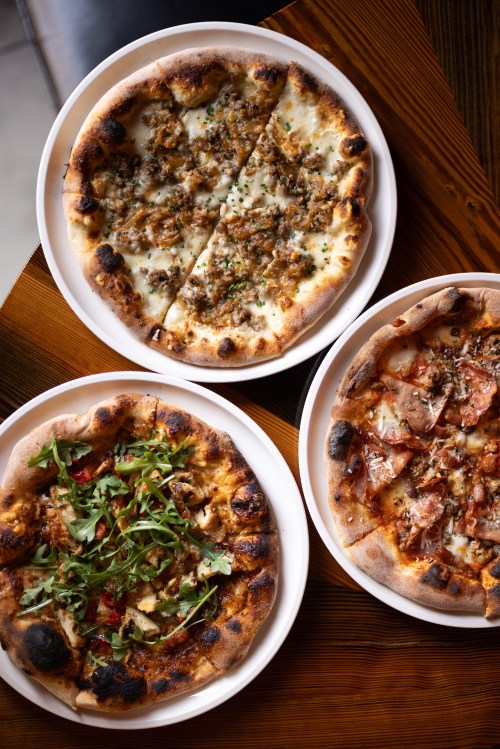 Three pizzas on white plates: one with meat, one with arugula, and one with various toppings, all on a wooden table.