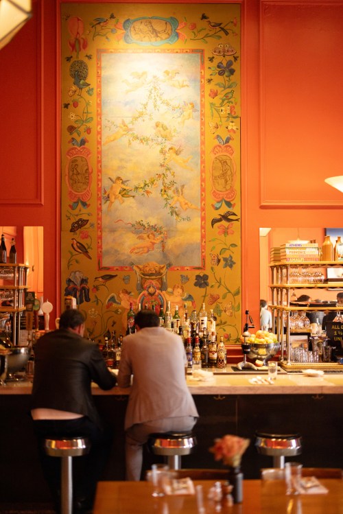 Two people sit at a bar, facing a colorful mural on the wall, surrounded by shelves of drinks and a cozy atmosphere.