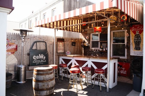 Outdoor bar with a striped awning, wooden barrels, and red stools, surrounded by vintage Coca-Cola signs.