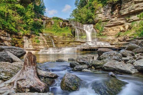 A serene waterfall cascades over rocky cliffs, surrounded by lush greenery and smooth stones in a tranquil river setting.