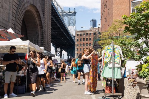 A bustling market scene under a bridge, with people browsing stalls and a woman examining colorful clothing.