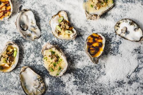 A variety of oysters on a textured surface, some topped with sauces and garnishes, others served raw.