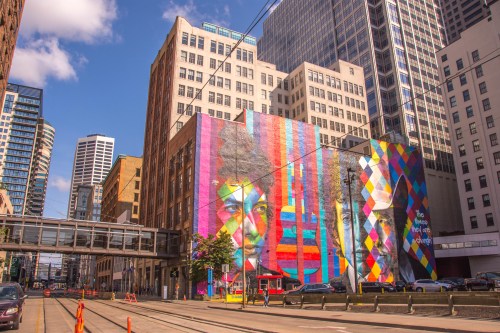 Colorful mural on a building in an urban setting, featuring abstract faces and vibrant patterns, with city skyscrapers in the background.