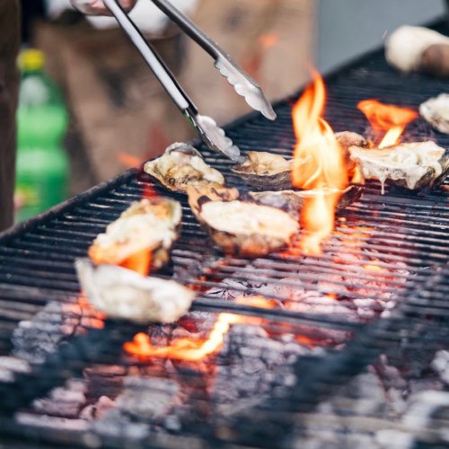 Grilled oysters on a barbecue grill with flames and tongs in action.
