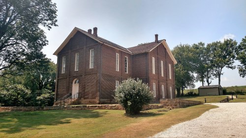 Historic brick building surrounded by greenery, with a gravel path leading up to it under a clear blue sky.