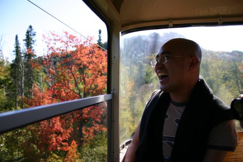 A smiling man sits inside a gondola, with vibrant autumn trees visible through the window.