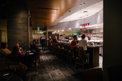 A modern restaurant interior with a bar, kitchen in the background, and diners enjoying their meals at tables.