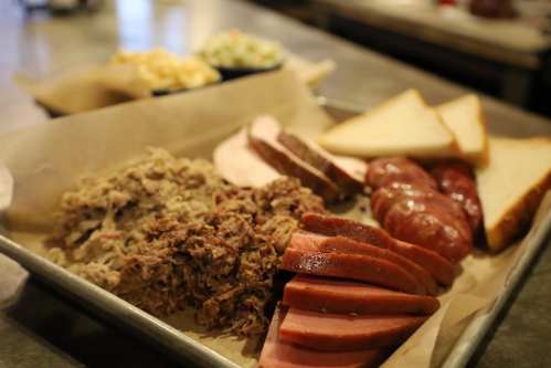 A platter of assorted meats including pulled pork, sliced sausage, and brisket, served with bread and sides.