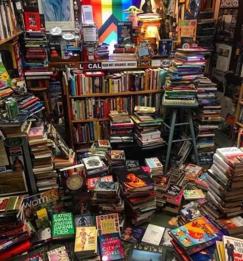 A cozy bookstore filled with stacks of books, colorful displays, and a pride flag in the background.