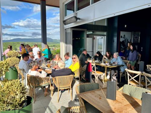 A lively outdoor restaurant scene with people dining, enjoying drinks, and a scenic view of mountains and clouds.