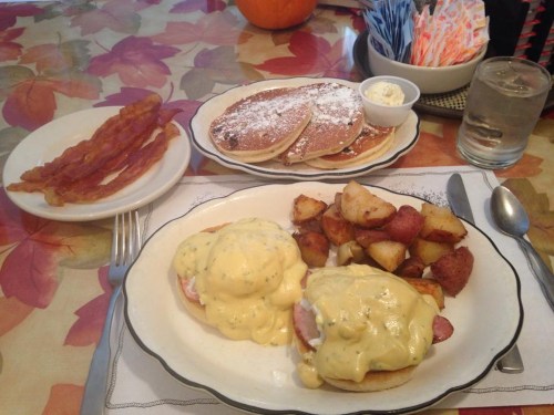 A plate of eggs Benedict with potatoes, bacon, and a side of pancakes with whipped cream on a colorful tablecloth.