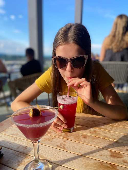 A young woman in sunglasses sips a drink at a table with two colorful cocktails, enjoying a sunny day.