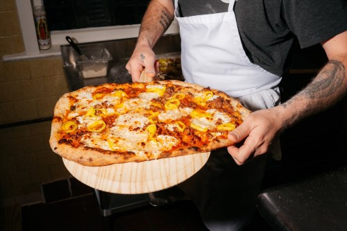 A person in an apron holds a freshly baked pizza topped with cheese and yellow peppers on a wooden pizza peel.