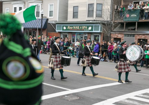 A St. Patrick's Day parade with drummers in kilts marching down the street, surrounded by festive crowds.