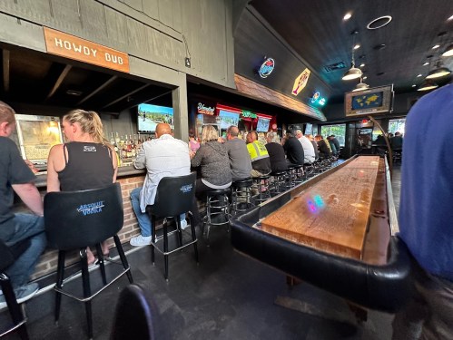 A lively bar scene with patrons seated at the counter, colorful lights, and a long wooden table in the foreground.