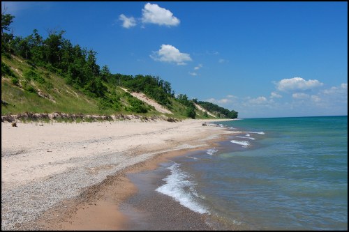 A serene beach scene with gentle waves, sandy shore, and lush green cliffs under a bright blue sky with fluffy clouds.