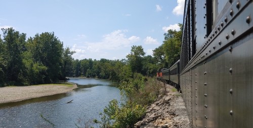 A train travels alongside a river, surrounded by lush greenery and a clear blue sky.