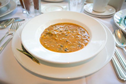 A bowl of thick soup with vegetables, served on a white plate, surrounded by utensils and glasses on a dining table.