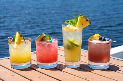 Four colorful cocktails on a wooden table by the water, featuring fruits and garnishes.