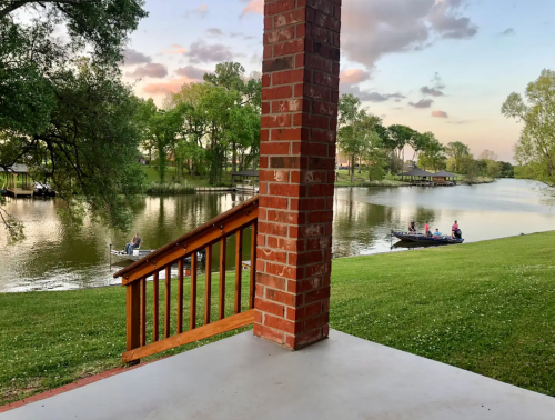 A serene lakeside view from a porch, with boats on the water and trees lining the shore under a colorful sky.