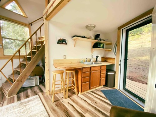 Cozy cabin interior featuring a small kitchen, wooden stairs, and large windows with natural light.