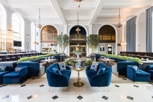 Elegant restaurant interior featuring blue velvet seating, greenery, and large windows, creating a bright, inviting atmosphere.