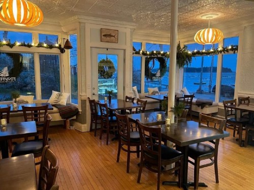 Cozy restaurant interior with wooden tables, warm lighting, and large windows overlooking a scenic waterfront.
