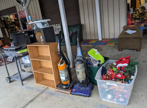A collection of items for sale, including vacuums, a wooden shelf, and a bin of holiday decorations outside a building.
