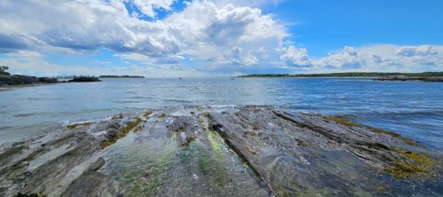 A serene coastal scene with rocky shorelines, gentle waves, and a blue sky dotted with clouds.