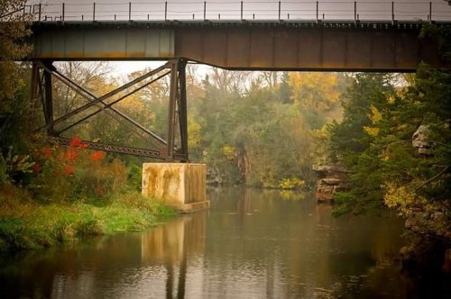 A rusted railway bridge spans a calm river, surrounded by trees with autumn foliage reflecting in the water.