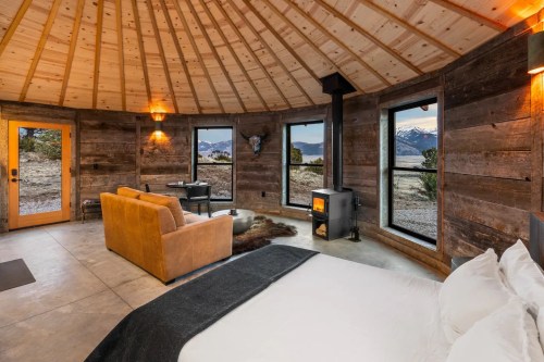 Cozy circular cabin interior with wooden walls, a bed, sofa, and large windows showcasing mountain views.