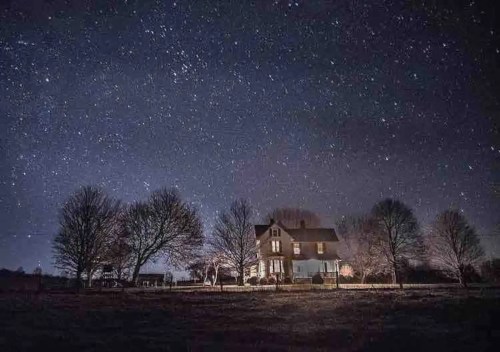 A house illuminated at night under a starry sky, surrounded by bare trees and a peaceful landscape.