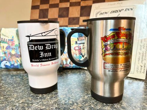 Two coffee mugs, one white with "Dew Drop Inn" and one stainless steel, on a countertop with a colorful background.