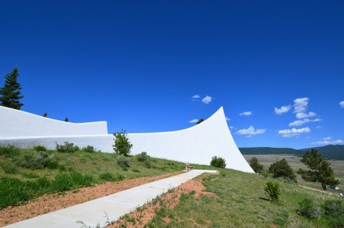 A modern white structure curves against a blue sky, surrounded by green grass and distant mountains.