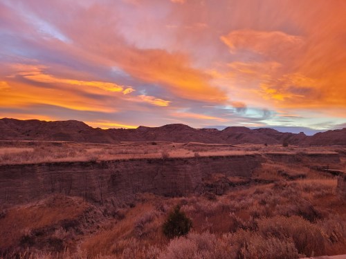 Vibrant sunset over rugged hills and dry grasslands, with colorful clouds illuminating the sky.