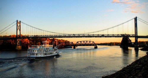 A riverboat glides along a calm river beneath two bridges at sunset, with trees lining the shore.