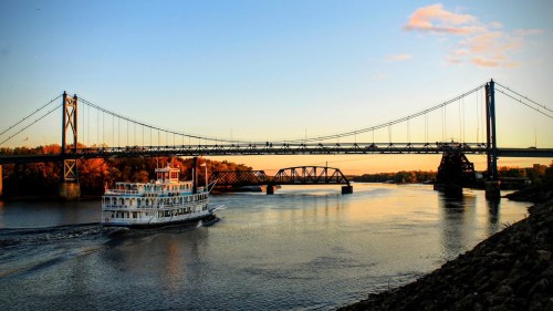 A riverboat sails under a bridge at sunset, reflecting warm colors on the water's surface.