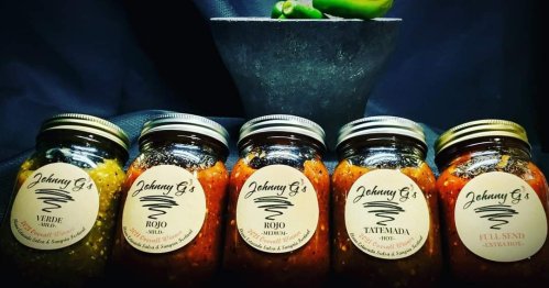Five jars of salsa labeled "Johnny G's" in various flavors: Verde, Rojo, Tatemada, and Full Send, with a bowl of peppers.