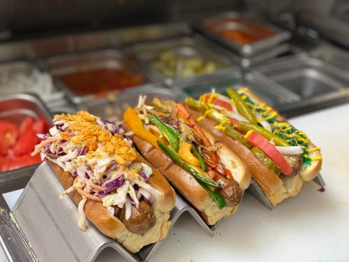 Four gourmet hot dogs topped with colorful vegetables and slaw, displayed on a counter with condiments in the background.