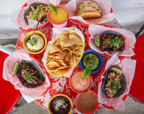 A colorful spread of tacos, chips, and dips on a red table, featuring various toppings and garnishes.