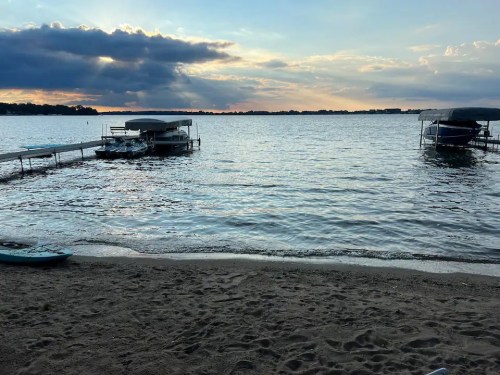 A serene lakeside view at sunset, featuring calm waters, sandy shore, and boat docks under a cloudy sky.