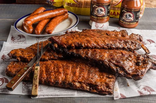 A platter of BBQ ribs and sausages with sauce bottles in the background, served on butcher paper.