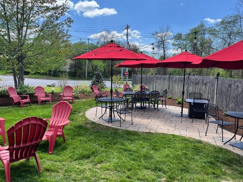 Outdoor seating area with red umbrellas and chairs on a sunny day, surrounded by greenery and a wooden fence.