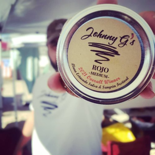 A person holds a jar labeled "Johnny G's Rojo - Medium" with a "2021 Overall Winner" sticker in a market setting.