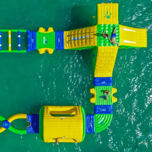 Aerial view of a colorful inflatable water park with people playing on the structures in a turquoise sea.