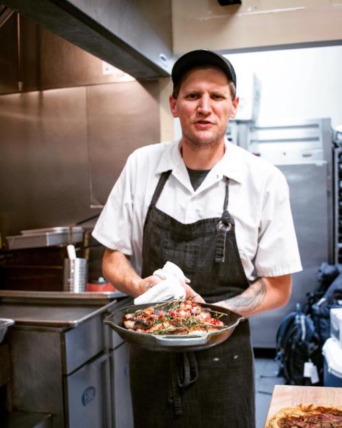 A chef in a kitchen holds a plate of grilled food, wearing an apron and cap, with a focused expression.