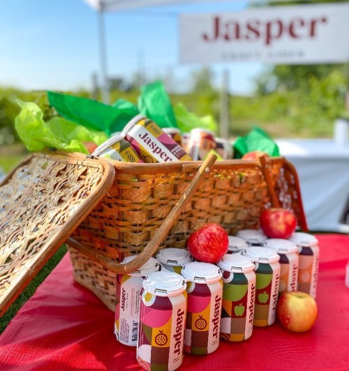 A picnic basket filled with colorful cans of Jasper cider and fresh apples on a red tablecloth outdoors.