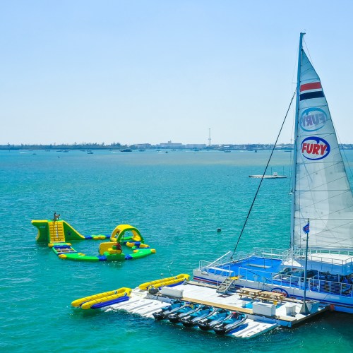 A catamaran with a sail docked near a colorful inflatable water park in a bright blue ocean.